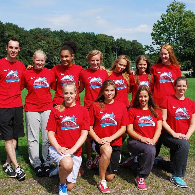 Gruppe von zehn Sportlerinnen und einem Trainer in roten T-Shirts, stehend auf einem Fußballplatz mit Bäumen im Hintergrund.
