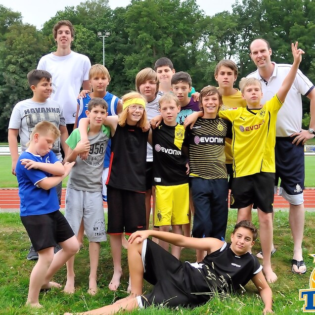 Gruppe von Jugendlichen in Sportkleidung posiert auf einem Rasen vor einem Sportplatz, einige lachen und zeigen V-Zeichen.