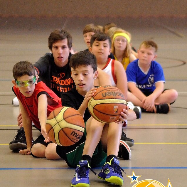 Gruppe von Kindern mit Basketballsitzen auf dem Boden, w&auml;hrend sie den Ball in Richtung Kamera rei&szlig;en.