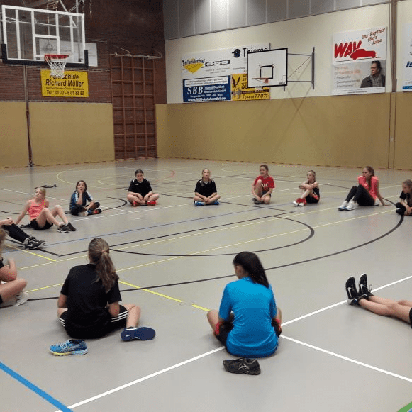 Mädchen sitzen im Kreis auf dem Boden einer Sporthalle während eines Trainings mit einer Trainerin.