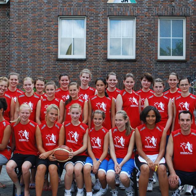 Gruppenshot von 20 Mädchen und einem Trainer in roten Sport-T-Shirts, die Basketball und ein Schulgebäude im Hintergrund zeigen.