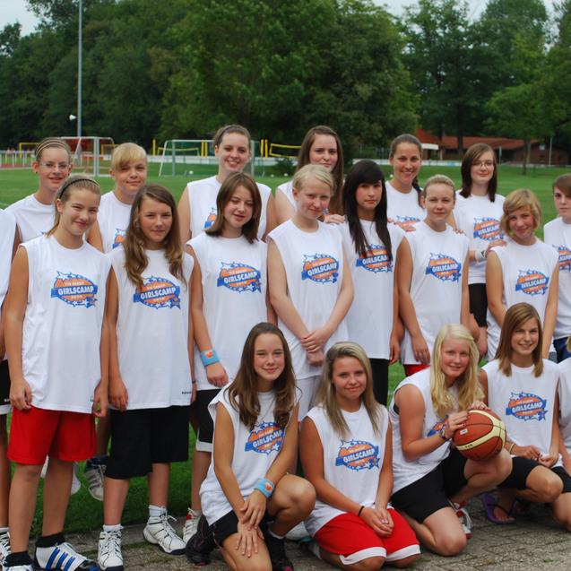 Gruppe von Mädchen in Trikots auf einem Sportplatz, einige halten Basketball, Lächeln und freundliche Atmosphäre.