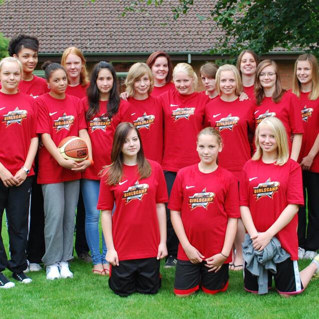 Gruppenfoto eines Basketballteams mit 15 Spielerinnen und 2 Trainern in roten T-Shirts, auf einer grünen Wiese.