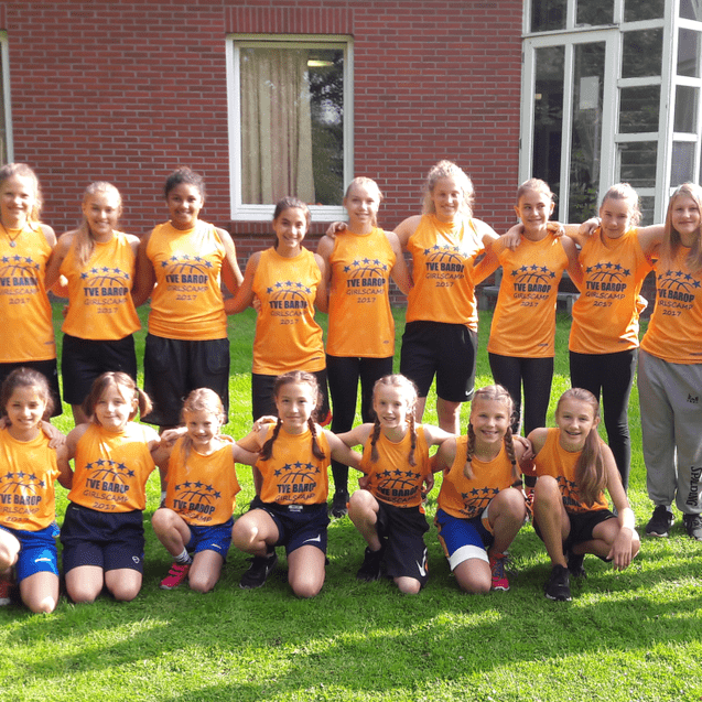 Gruppe junger Sportlerinnen in orangefarbener Teambekleidung posiert lächelnd auf einer Wiese vor einem Gebäude.