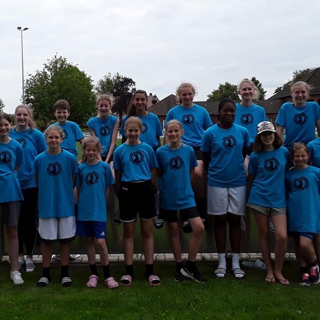 Gruppe von 19 Personen in blauen T-Shirts steht lächelnd vor einem Fußballfeld mit grünem Rasen.