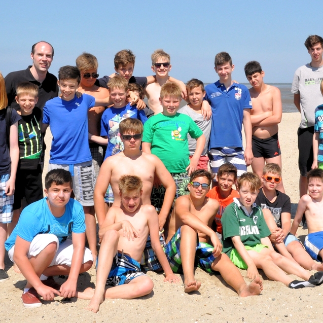 Gruppe von Jungen und Erwachsenen mit Badesachen stehen am Strand, l&auml;chelnd vor dem Meer. Sonniger Himmel im Hintergrund.