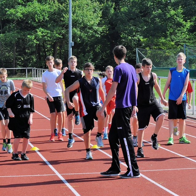 Gruppe von Jugendlichen in Sportkleidung beim Aufw&auml;rmen auf einer Tartanbahn, angeleitet von einem Trainer.