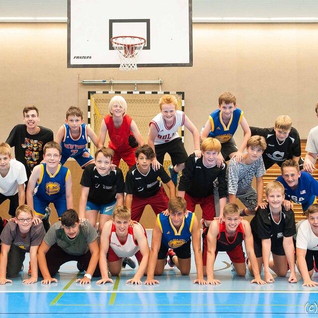Gruppenshooting von Jungen in Basketballtrikots, die auf dem Boden gestapelt stehen, in einer Sporthalle.
