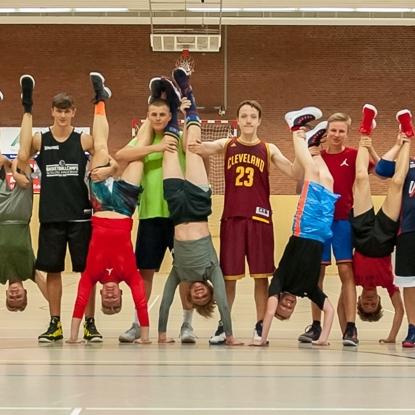 Gruppierung von Jugendlichen in Sportkleidung, viele davon stehen auf Händen, in einer Sporthalle.