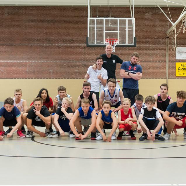Gruppe von Basketballspielern in einer Sporthalle, posierend in zwei Reihen vor einem Basketballkorb.