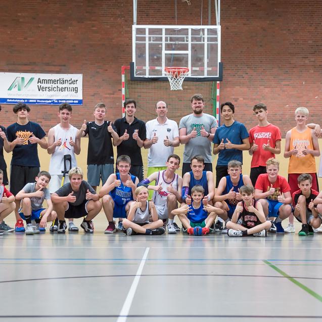 Gruppenfoto von Basketballspielern in einer Halle, alle zeigen Daumen hoch, Basketballkorb im Hintergrund.