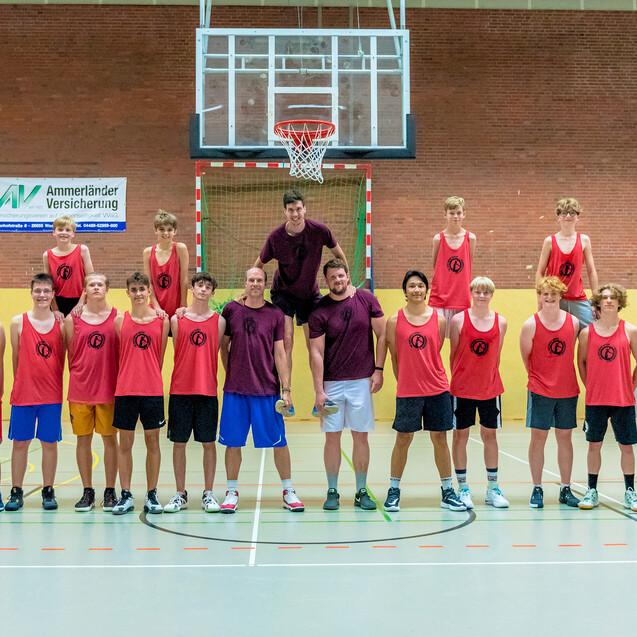 Gruppenfoto einer Basketballmannschaft in roten Trikots auf einem Feld mit Korb im Hintergrund.