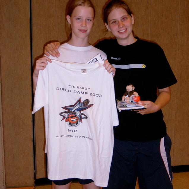 Zwei Mädchen in Sportkleidung halten ein T-Shirt mit Aufdruck und eine Trophäe in einer Sporthalle.