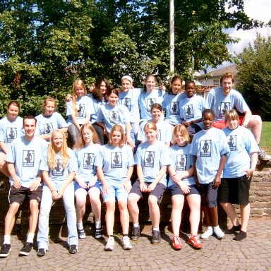 Gruppe von Jugendlichen in blauen T-Shirts, sitzend und stehend auf einer Steinmauer vor einem grünen Hintergrund.