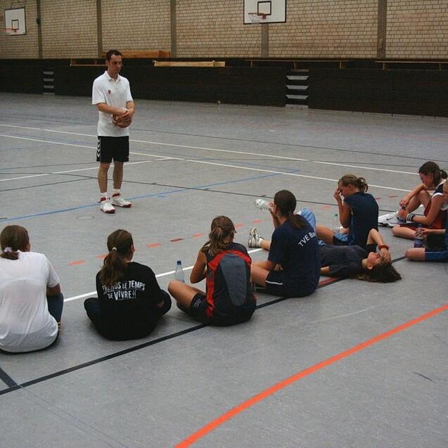 Trainer steht vor einer Gruppe sitzender Spielerinnen in einer Sporthalle während einer Basketballübung.