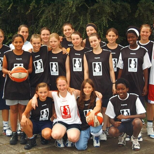 Gruppenaufnahme von 16 Mädchen in Basketballtrikots, einige halten einen Basketball, umgeben von Bäumen.
