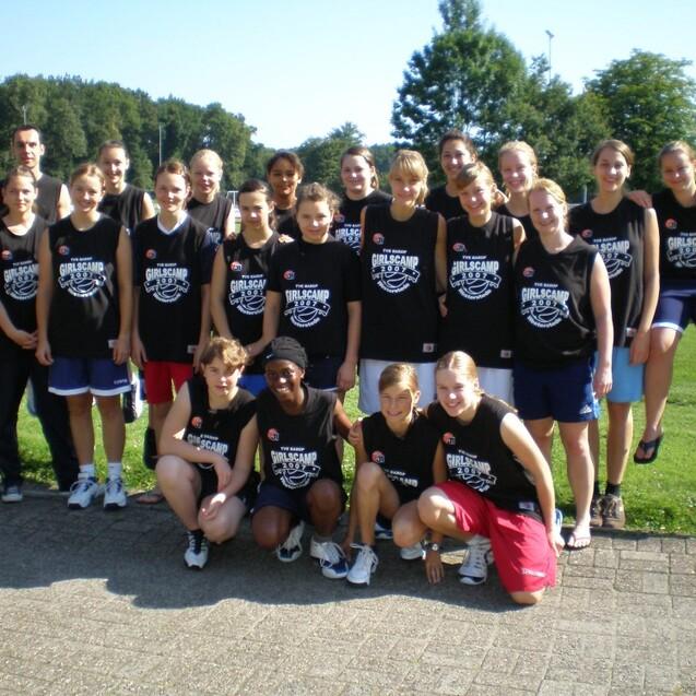 Gruppe von Mädchen in Basketballtrikots posiert lächelnd auf einem Sportplatz unter blauem Himmel.