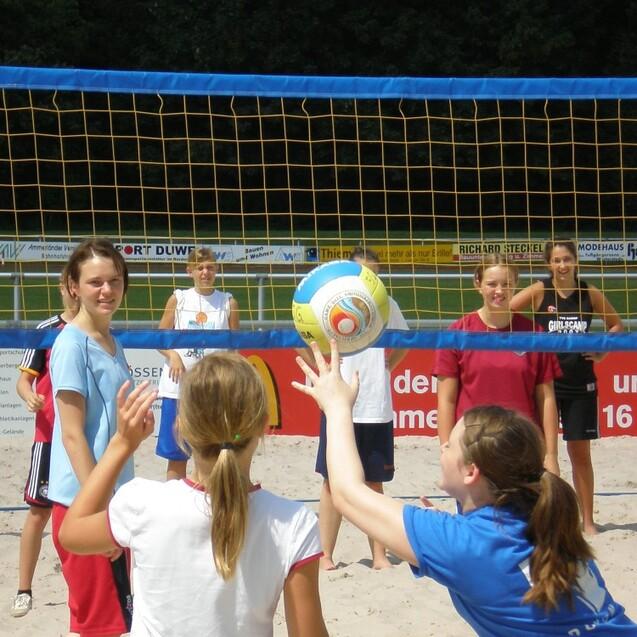 Gruppe von Jugendlichen spielt Beachvolleyball im Sand unter einem Netz, während der Ball gerade in die Luft geworfen wird.