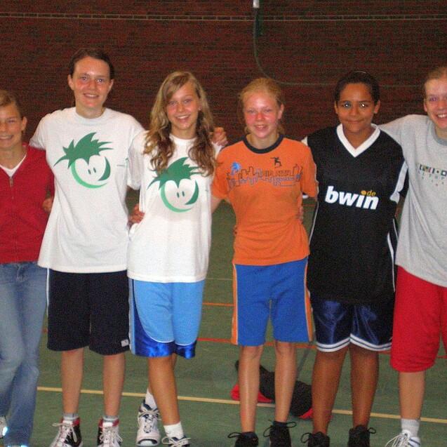 Sechs Mädchen in Sportkleidung stehen lächelnd auf einem Basketballplatz nebeneinander.
