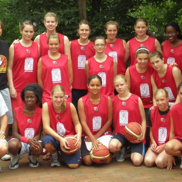Gruppenfoto einer Basketballmannschaft mit 16 Spielerinnen und 2 Trainern, alle in roten Trikots, auf einem Außenfeld.