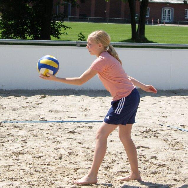 Mädchen mit blonden Haaren spielt Beachvolleyball, ball wirbelt in der Luft vor ihr auf einem Sandplatz.