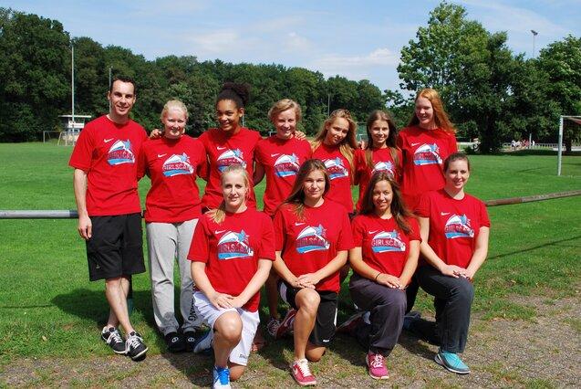 Gruppe von zehn Sportlerinnen und einem Trainer in roten T-Shirts, stehend auf einem Fußballplatz mit Bäumen im Hintergrund.