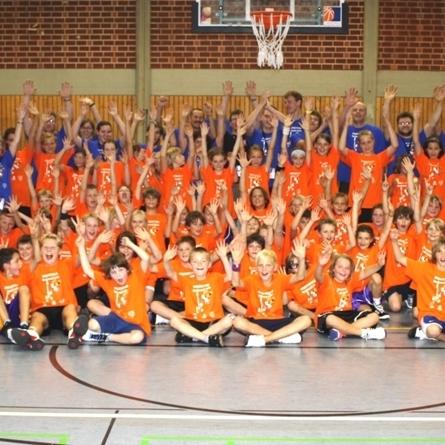 Gruppenshot von Kindern und Erwachsenen in orangefarbenen und blauen T-Shirts, die in einer Sporthalle jubeln.