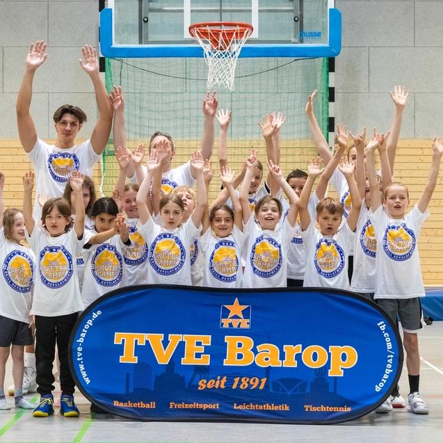 Gruppe von Kindern und Jugendlichen in T-Shirts, die begeistert vor einem Basketballkorb stehen und Hände heben.