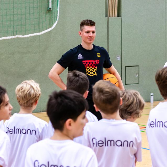 Trainer spricht mit einer Gruppe von Kindern in Basketball-T-Shirts in einer Sporthalle. Ein Junge hält einen Basketball.