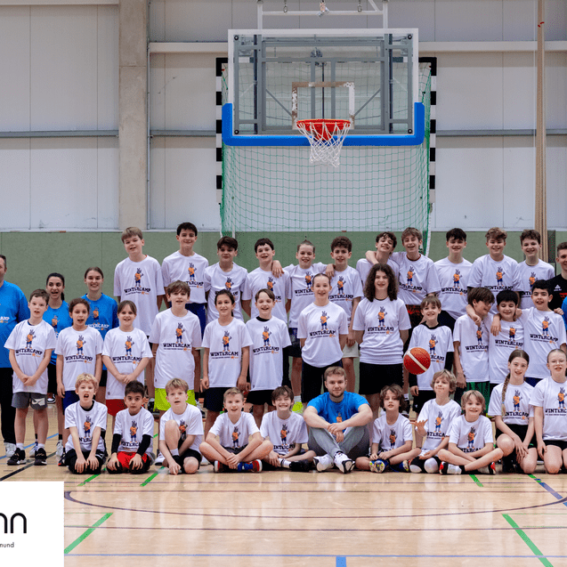 Gruppenfoto von Kindern und Trainern in Sportshirts in einer Halle, Basketballkorb und Sponsorenlogo im Vordergrund.