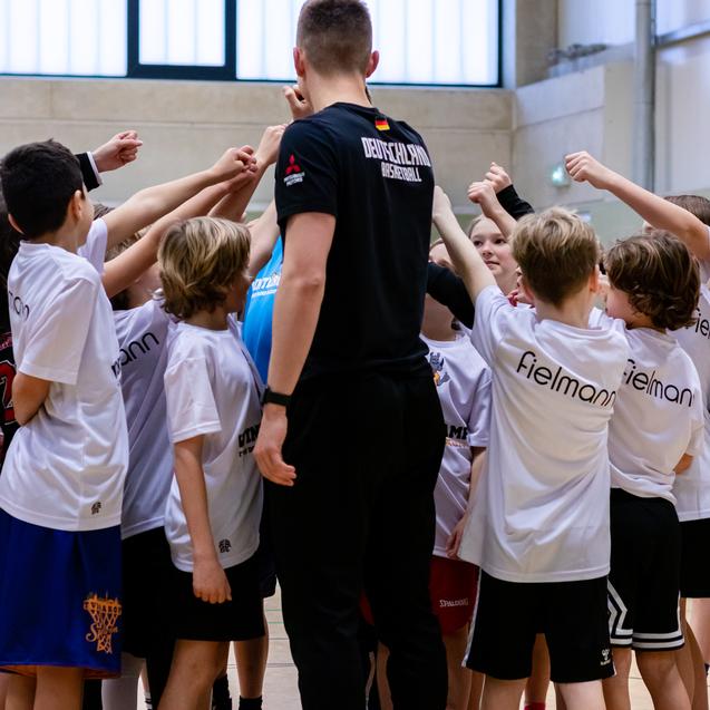 Gruppierung von Kindern in weißen T-Shirts, die gemeinsam die Hände in die Mitte eines Basketballspielers heben.