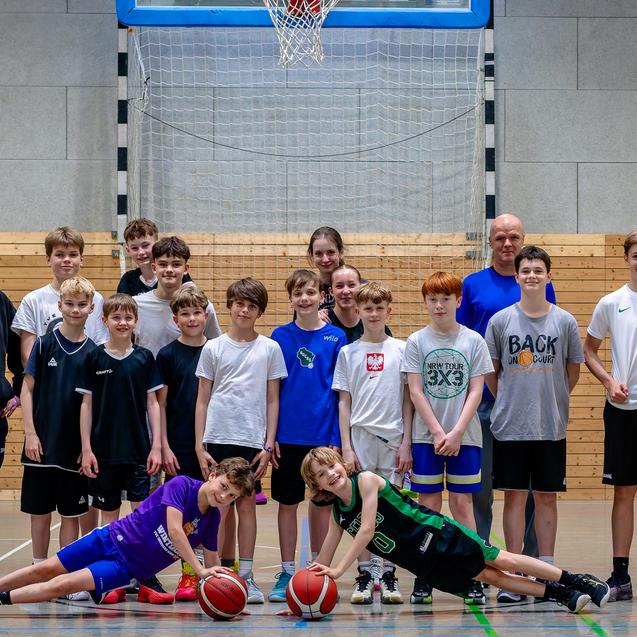 Gruppe von Jugendlichen in Sportkleidung, die auf einem Basketballfeld posieren, mit zwei Basketballs in der Mitte.