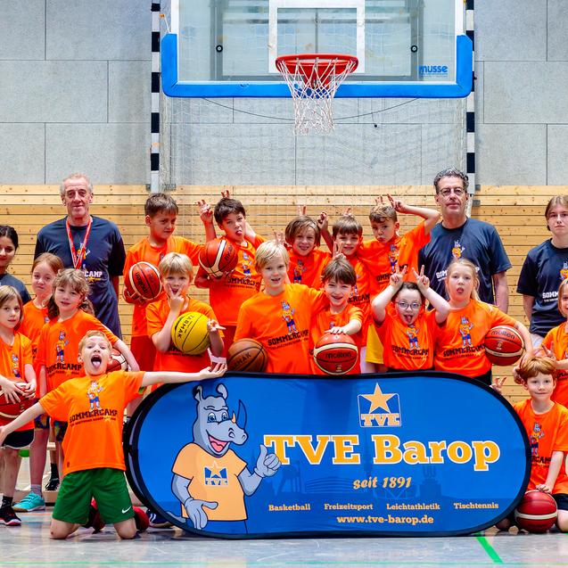 Gruppenbild von Kindern in orangenen T-Shirts mit Basketballs, die vor einem Basketballkorb und einem TVE Barop Banner stehen.