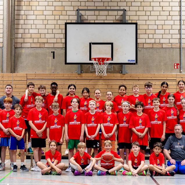Gruppenfoto einer Basketballmannschaft in roten Trikots, posesierend vor einem Basketballkorb in einer Sporthalle.