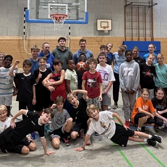Gruppe von Kindern und Jugendlichen posiert gemeinsam auf einem Basketballplatz mit Korb im Hintergrund.