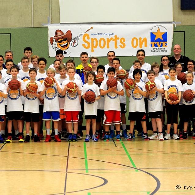 Gruppenfoto von Kindern und Trainern auf einem Basketballfeld, alle halten Basketballs, mit einem Banner im Hintergrund.