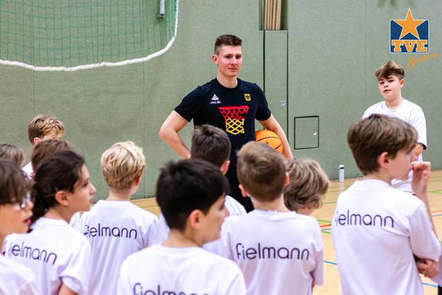 Trainer spricht mit einer Gruppe von Kindern in Basketball-T-Shirts in einer Sporthalle. Ein Junge hält einen Basketball.