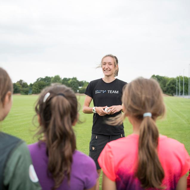 Trainerin spricht mit einer Gruppe von M&auml;dchen auf einem Fu&szlig;ballfeld unter bew&ouml;lktem Himmel.