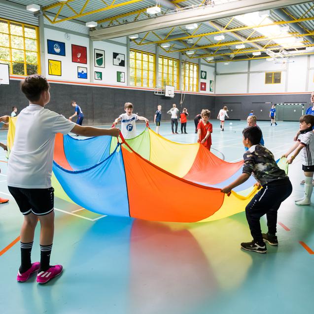 Kinder halten ein gro&szlig;es, buntes Fallschirmspielzeug in einer Sporthalle, w&auml;hrend sie miteinander spielen und lachen.