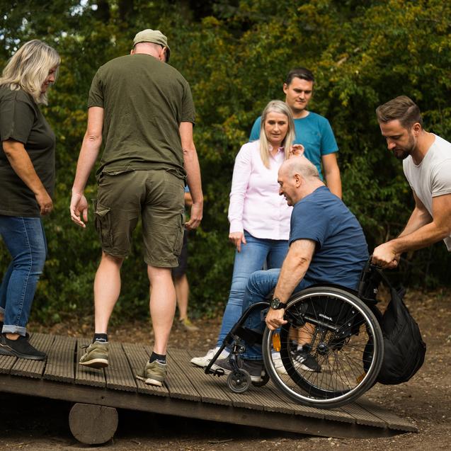 Gruppenszenen mit mehreren Personen, die einem Rollstuhlfahrer beim &Uuml;berwinden einer schr&auml;gen Holzrampe helfen.