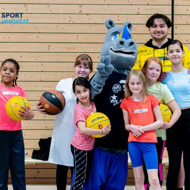 Gruppe von sechs Kindern und zwei Erwachsenen, darunter ein Maskottchen, posieren mit Sportbällen in einer Halle.