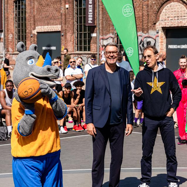 Ein Maskottchen in Tierkostüm hält einen Basketball, während zwei Männer auf einem Sportplatz stehen und zuschauen.