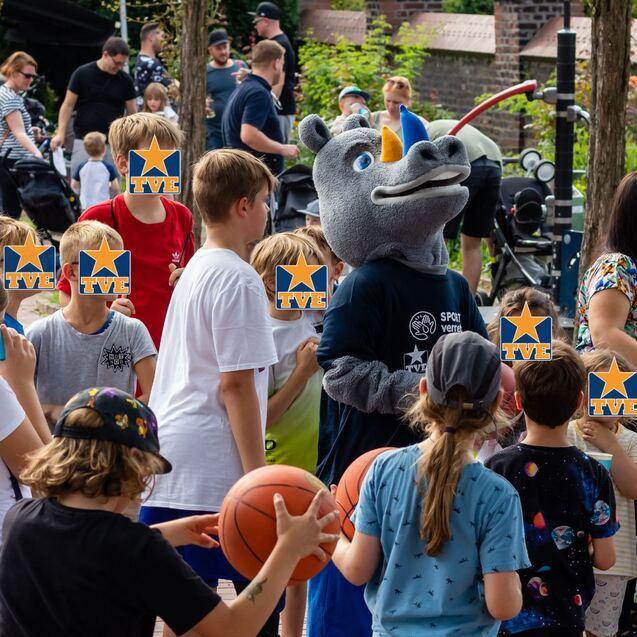 Kinder versammeln sich um ein Maskottchen in einem grauen Tierkostüm auf einem Fest, mit Basketball in der Mitte der Gruppe.