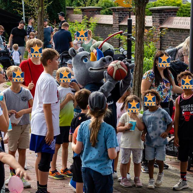 Eine Gruppe von Kindern umringt ein Maskottchen, das einen Basketball hält, in einer lebhaften Umgebung.