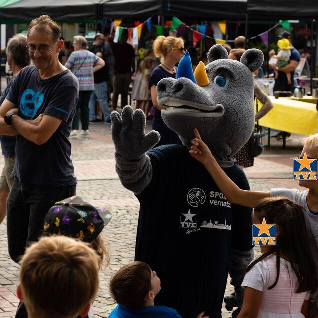 Ein Maskottchen in Rhinozeros-Kostüm winkt Kindern auf einem belebten Festplatz mit Ständen und bunten Dekorationen.