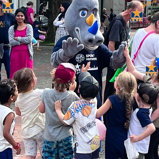 Eine animierte Figur in Rhino-Charakter spielt mit Kindern auf einem Fest, umgeben von Zuschauern.