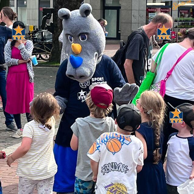 Maskottchen in tierischer Kostüm begrüßt Kinder auf einer Straße, während Passanten im Hintergrund stehen.