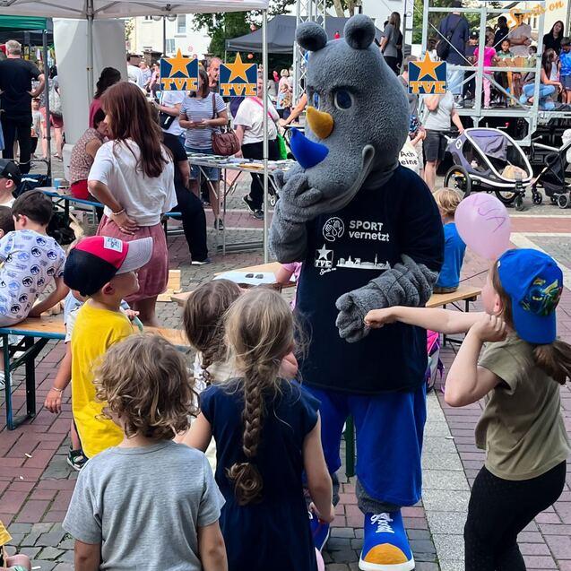 Maskottchen interagiert mit Kindern auf einer Veranstaltung, umgeben von Ständen und Zuschauern im Hintergrund.