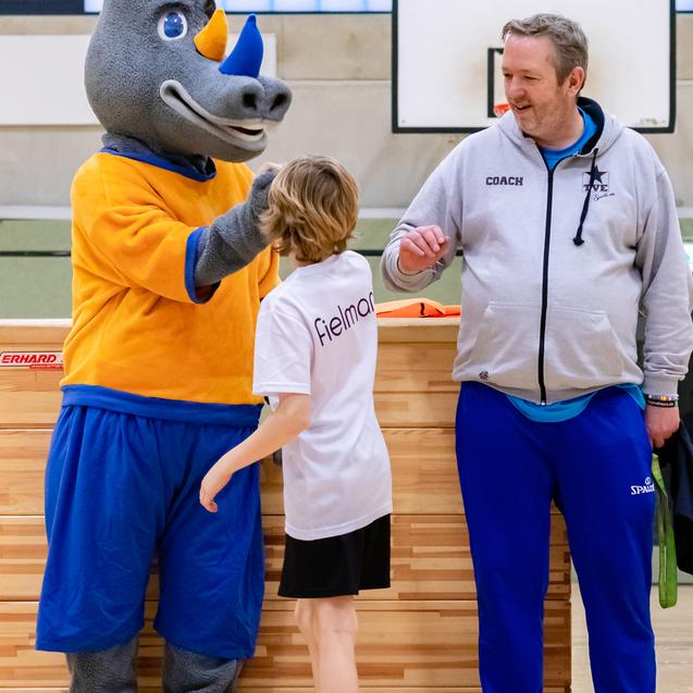 Maskottchen umarmt ein Kind in Sportkleidung, während ein Trainer daneben steht, in einer Sporthalle.