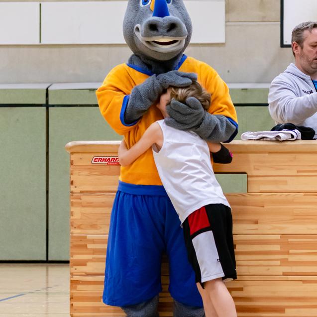 Ein Kind umarmt ein Maskottchen in einem sportlichen Outfit an einem Tisch in einer Sporthalle.